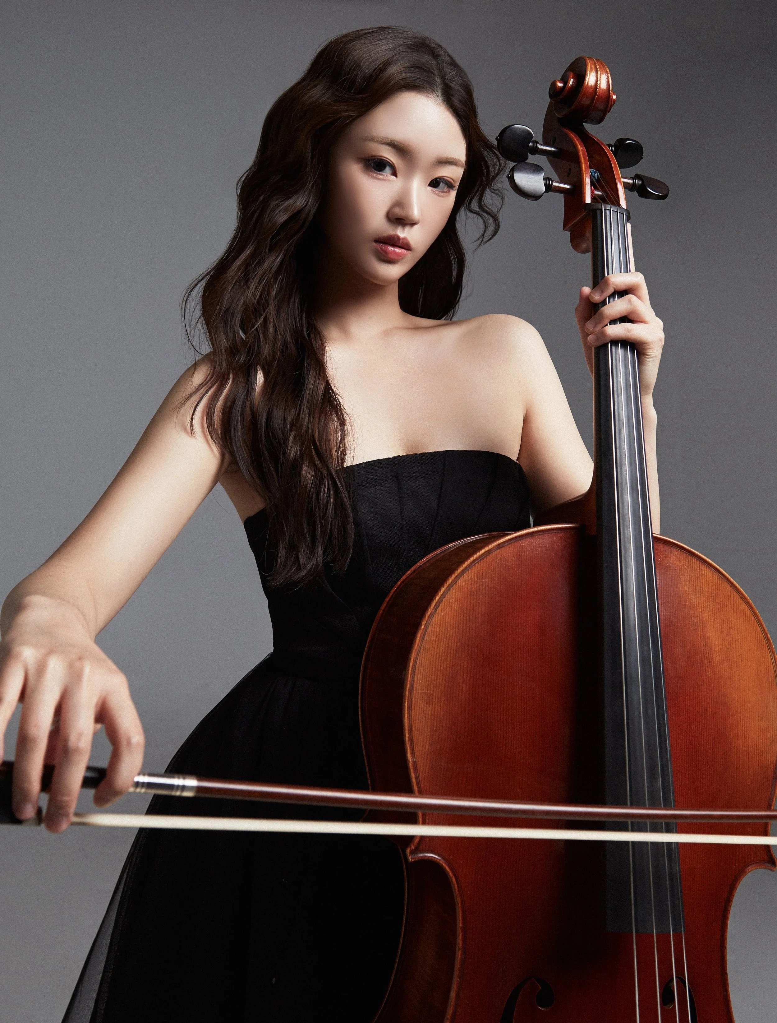 A woman in a black strapless dress playing a cello, holding a bow, with a neutral gray background.