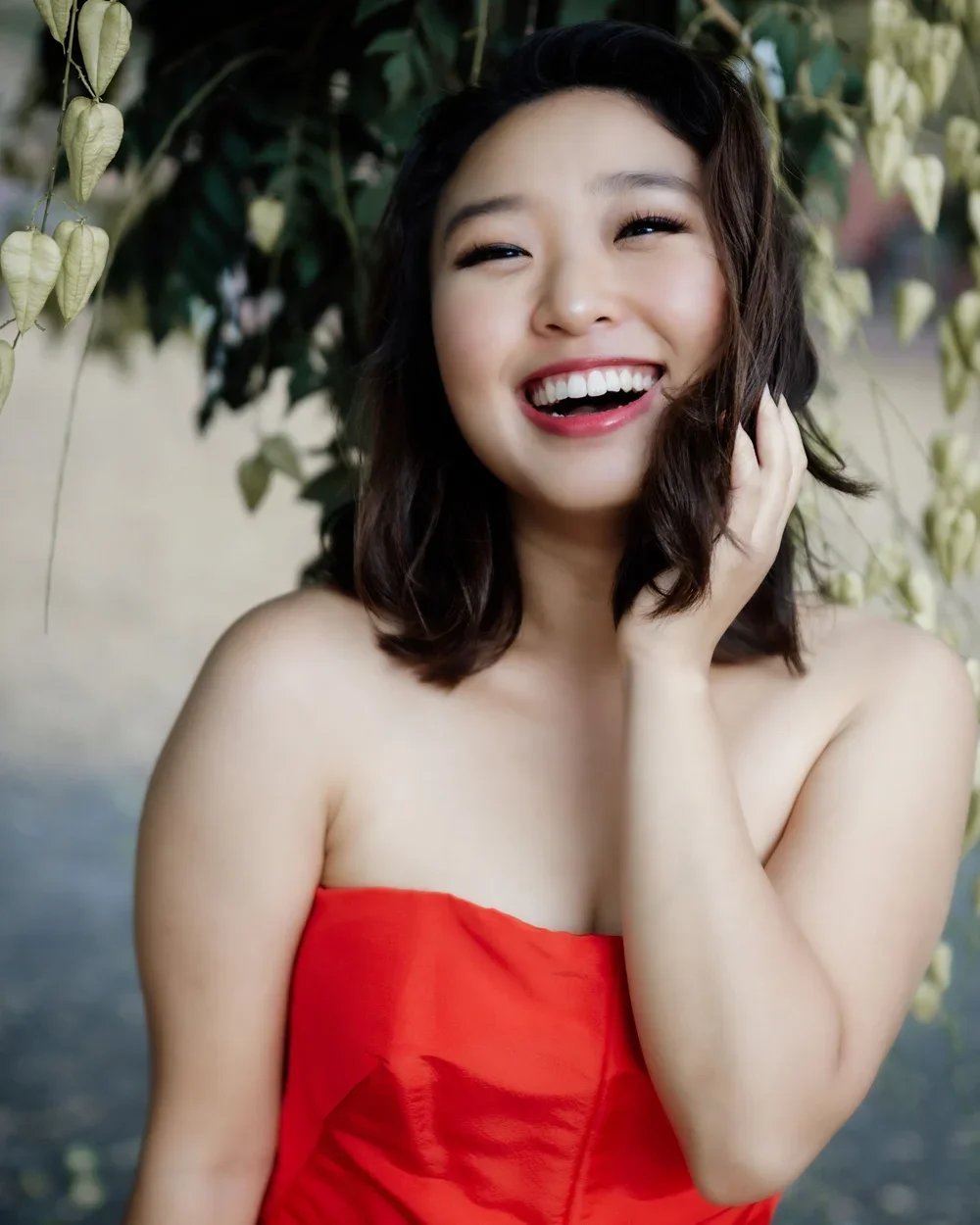 A woman with shoulder-length dark hair, smiling and touching her neck, wearing a strapless red dress, standing outdoors with green foliage in the background.