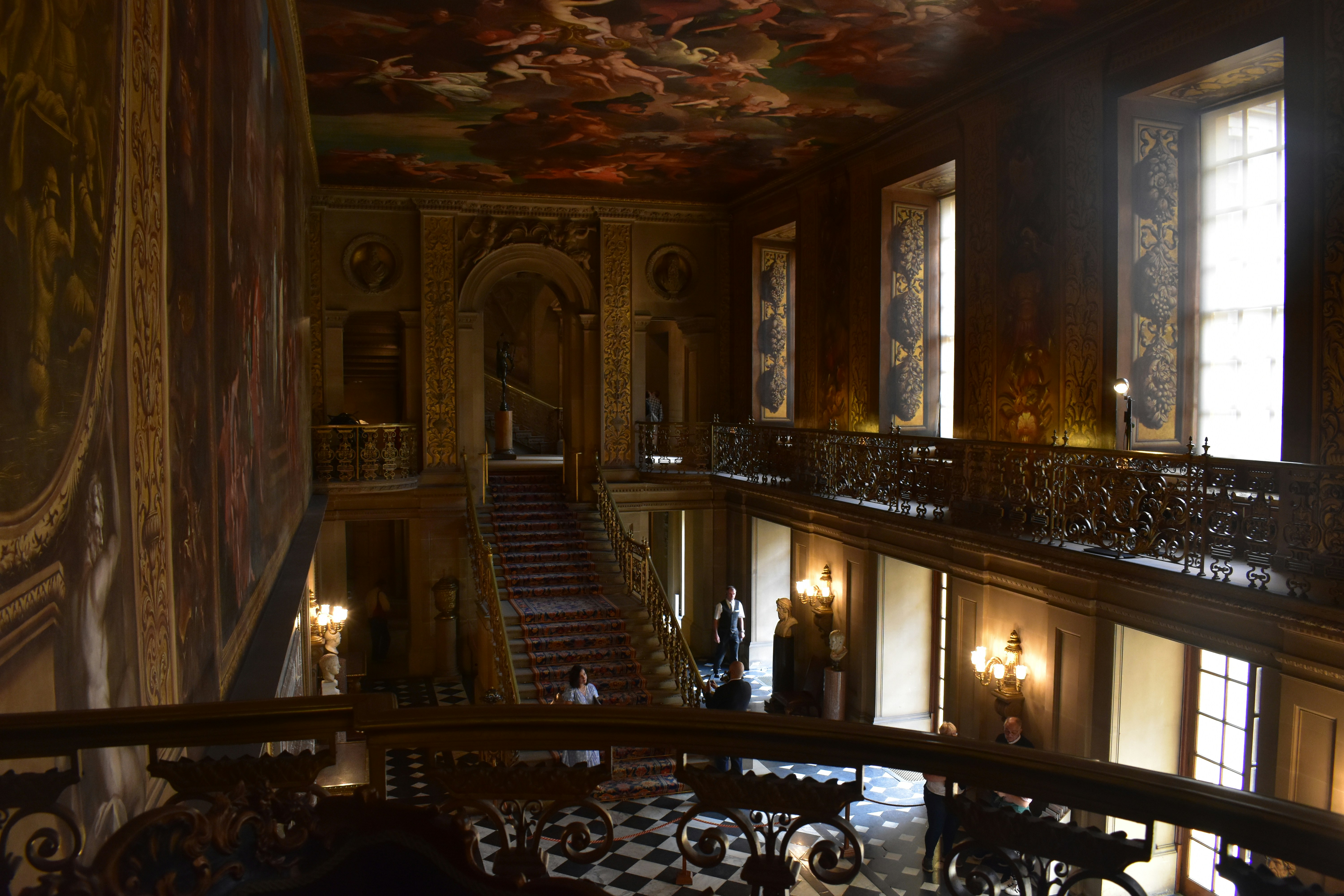 An ornate interior staircase beneath painted ceilings, with tall windows and decorative railings.