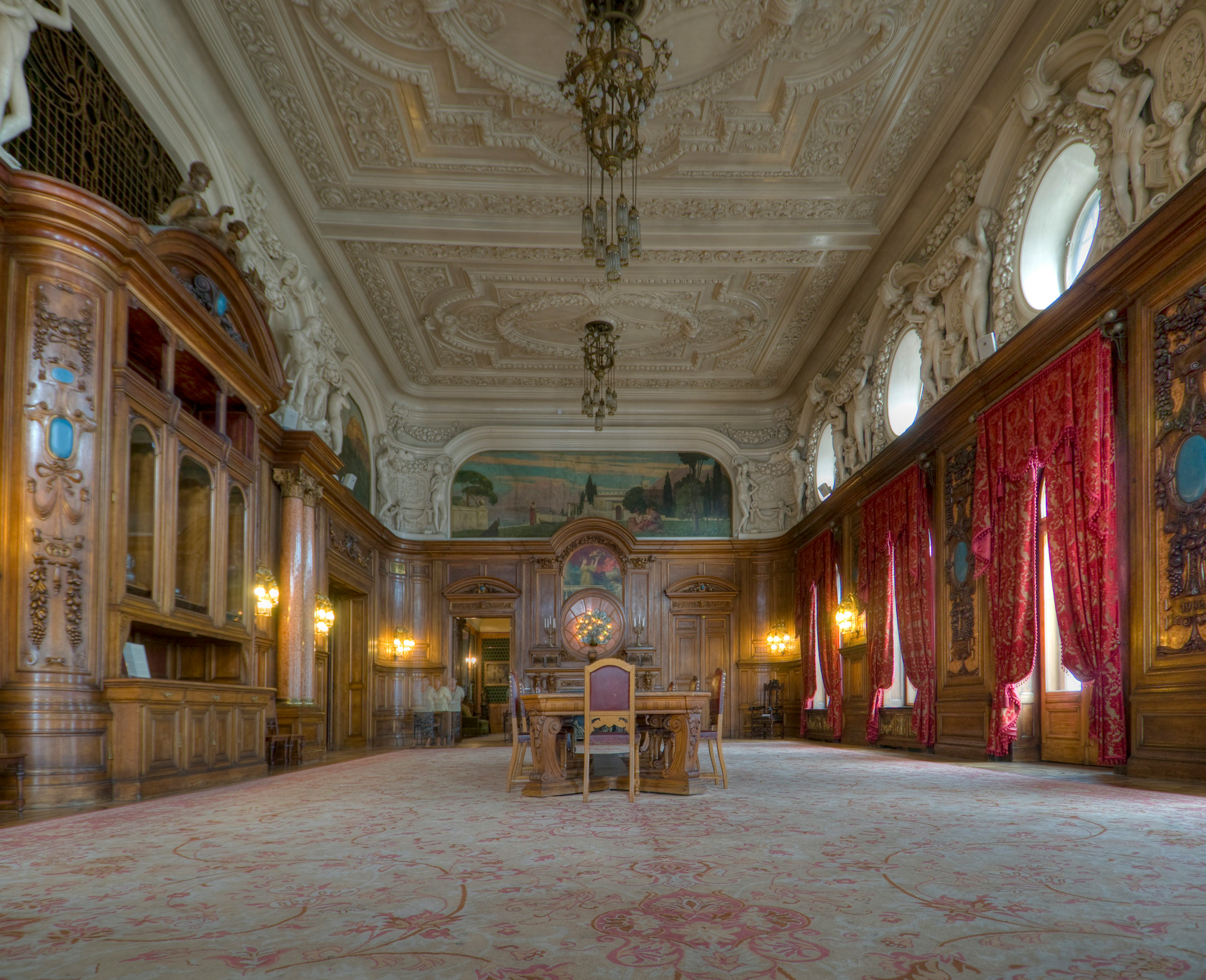 An ornate wood-paneled hall with a decorated ceiling, red curtains, and a central chair.