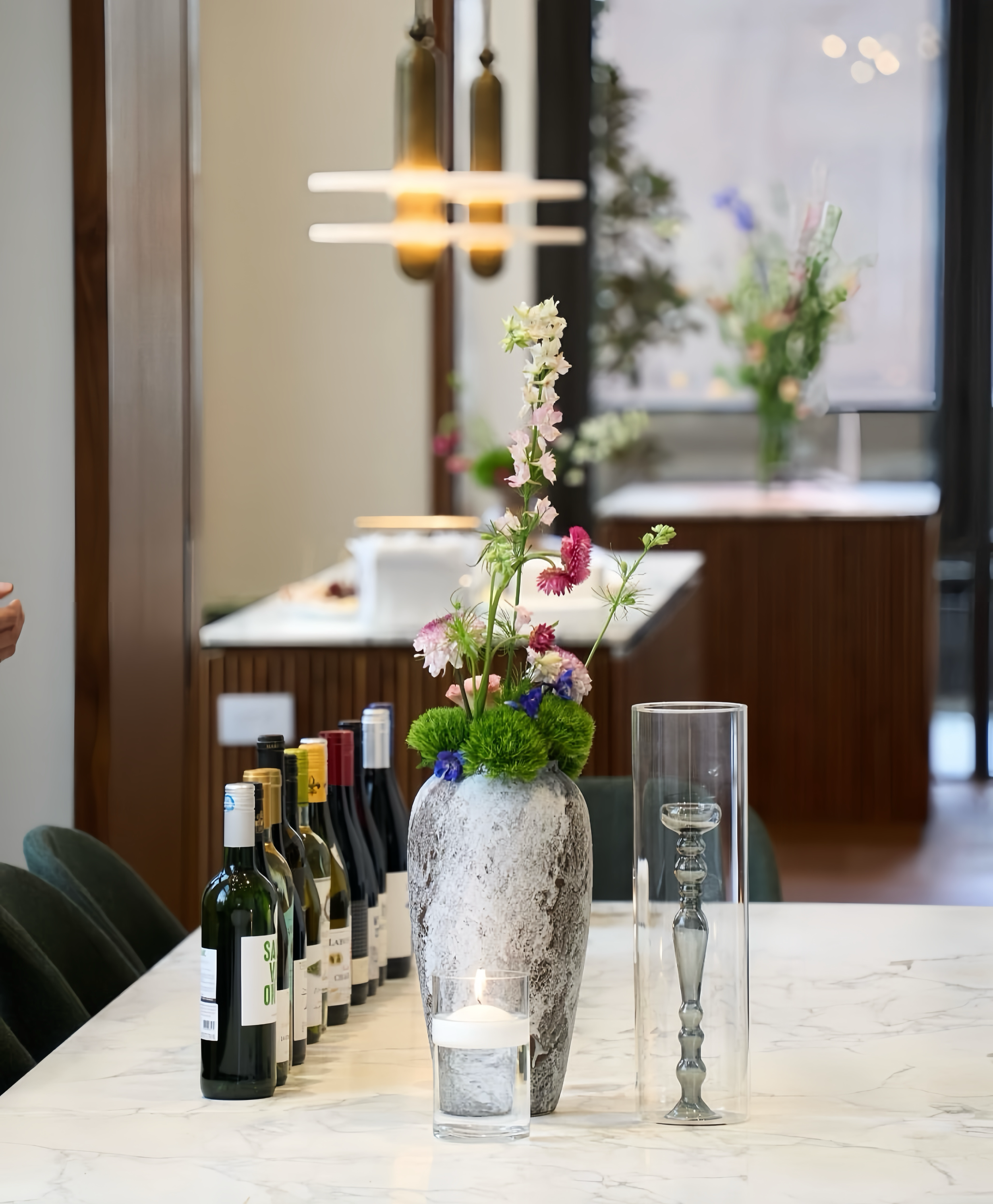 A decorative table setting with a large vase of pink, white, and purple flowers, several bottles of wine, a lit candle in a glass holder, a glass hurricane lamp, and additional vases of flowers in the background.