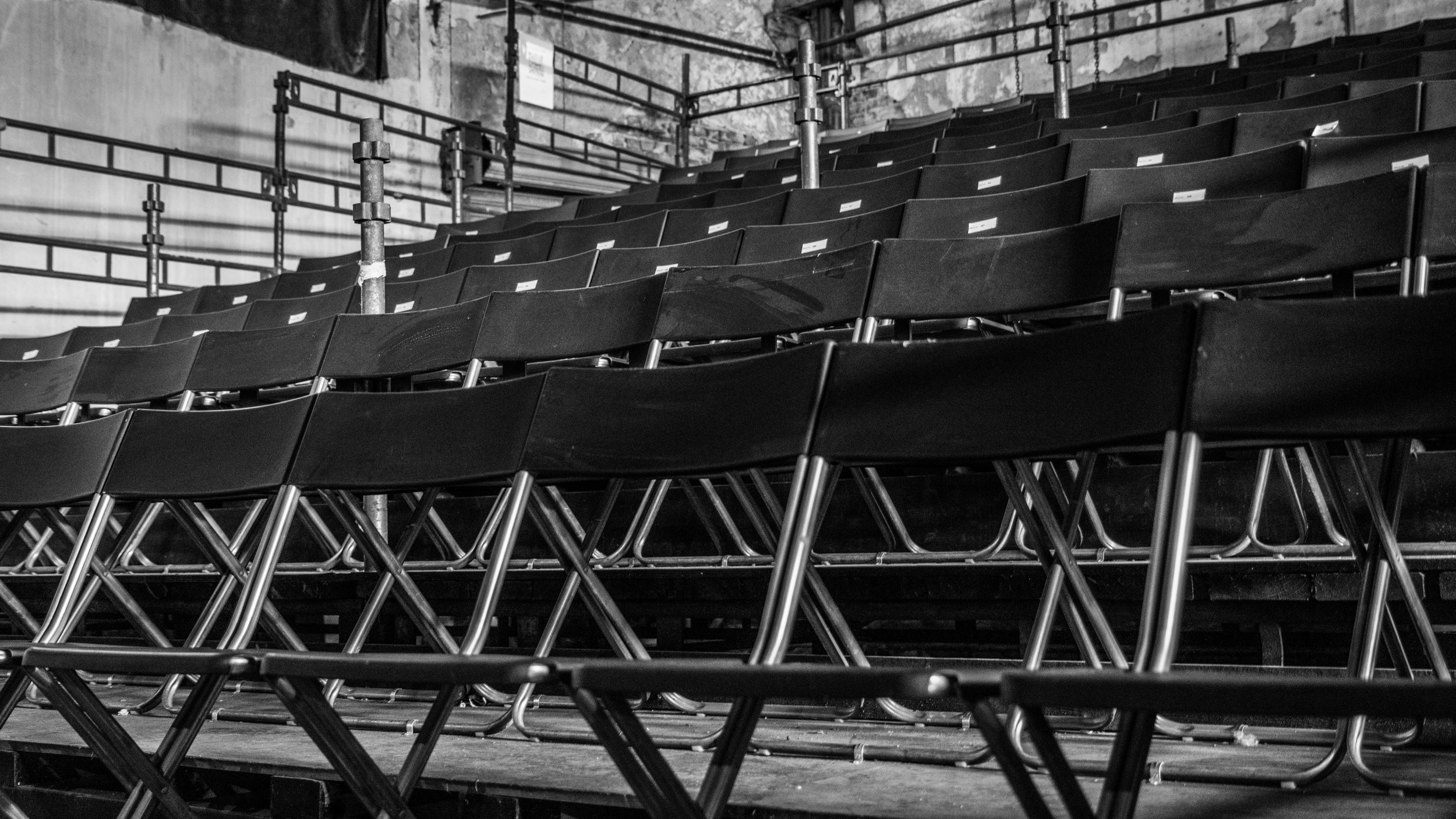 Empty black folding chairs in an indoor auditorium or theater with metal railings and an industrial brick wall in the background.
