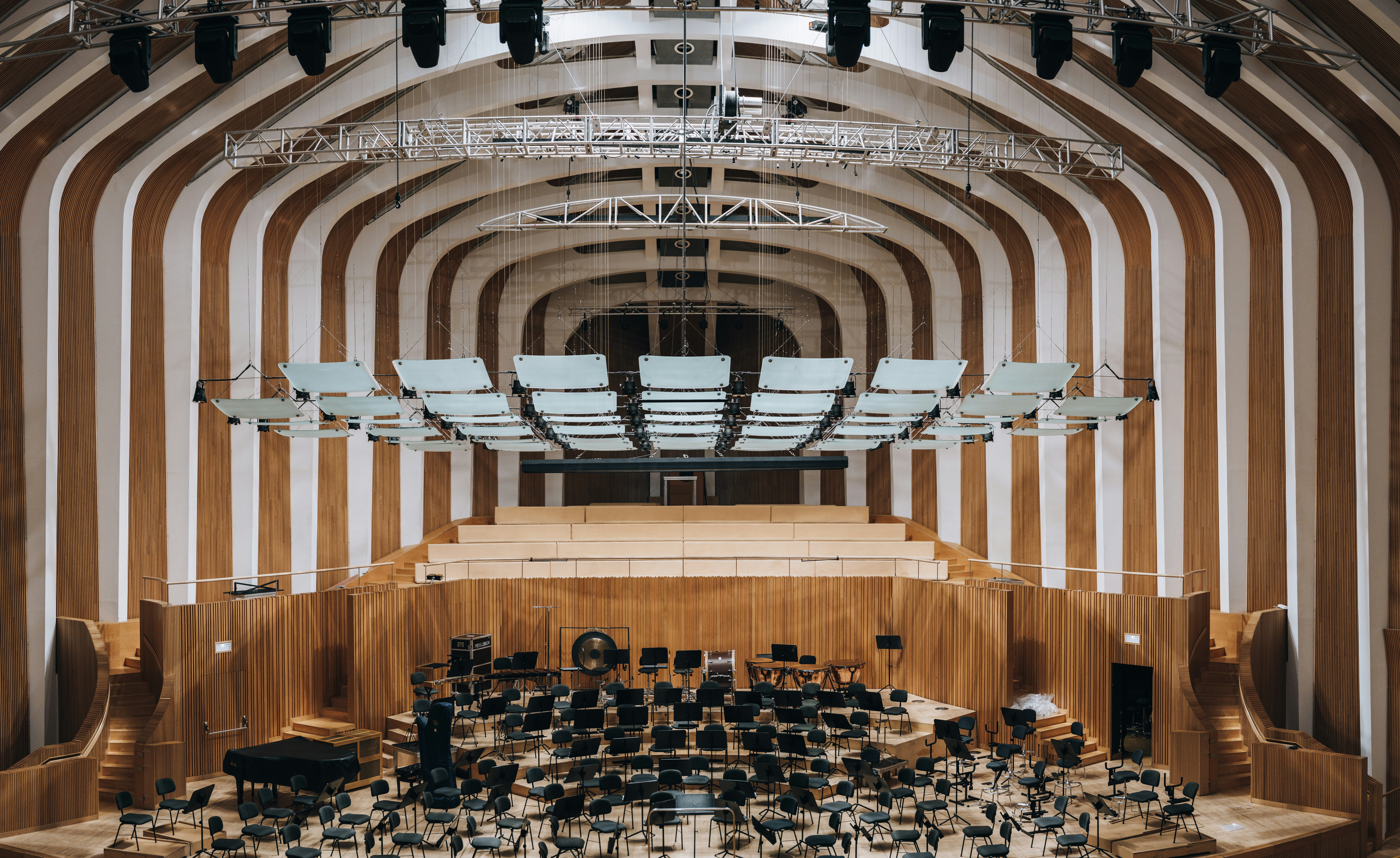 An empty concert hall stage with a grand piano, orchestral chairs and music stands beneath suspended acoustic panels.