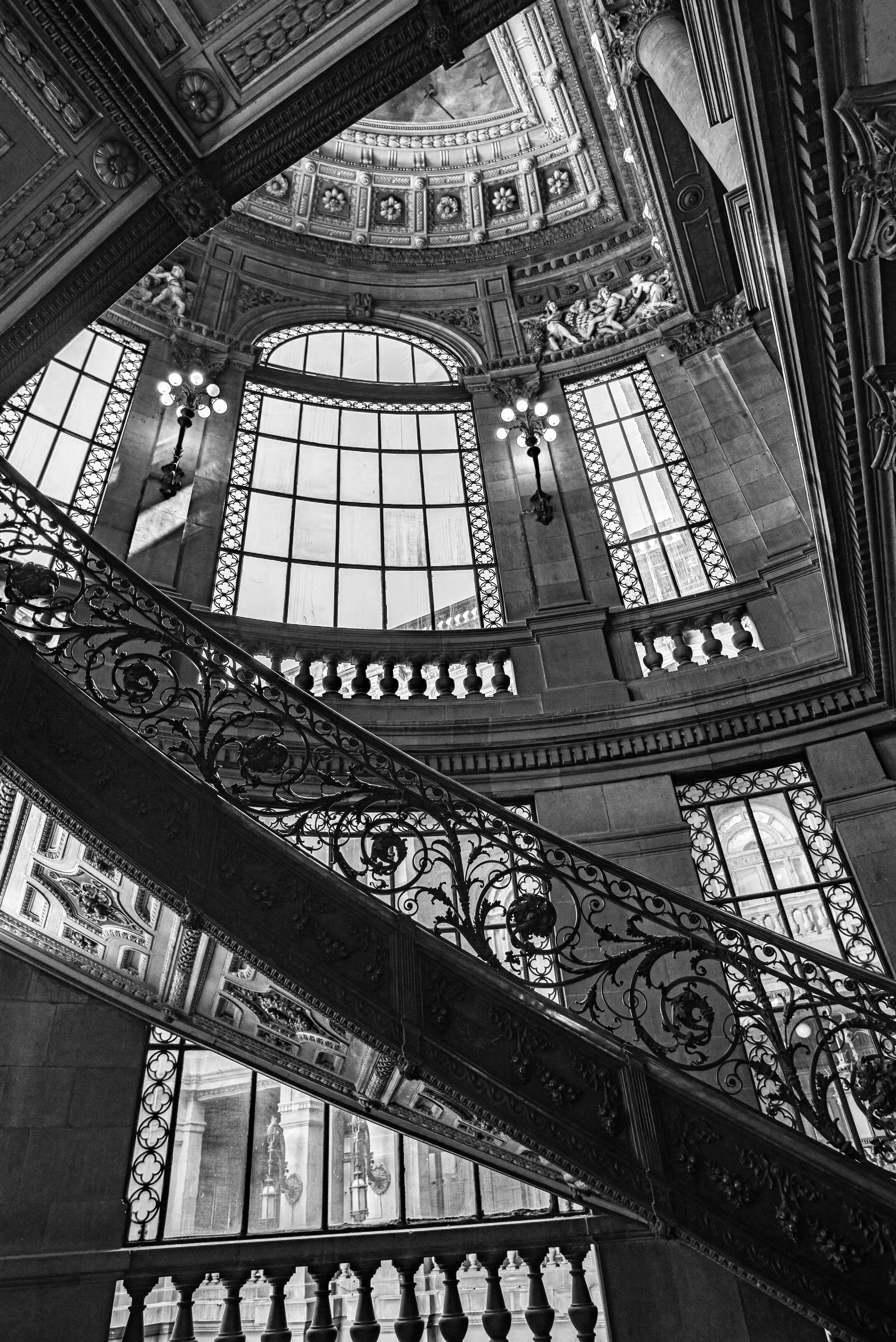 Interior view of a historic building with ornate architectural details, large windows, iron railing staircase, and decorative ceiling.