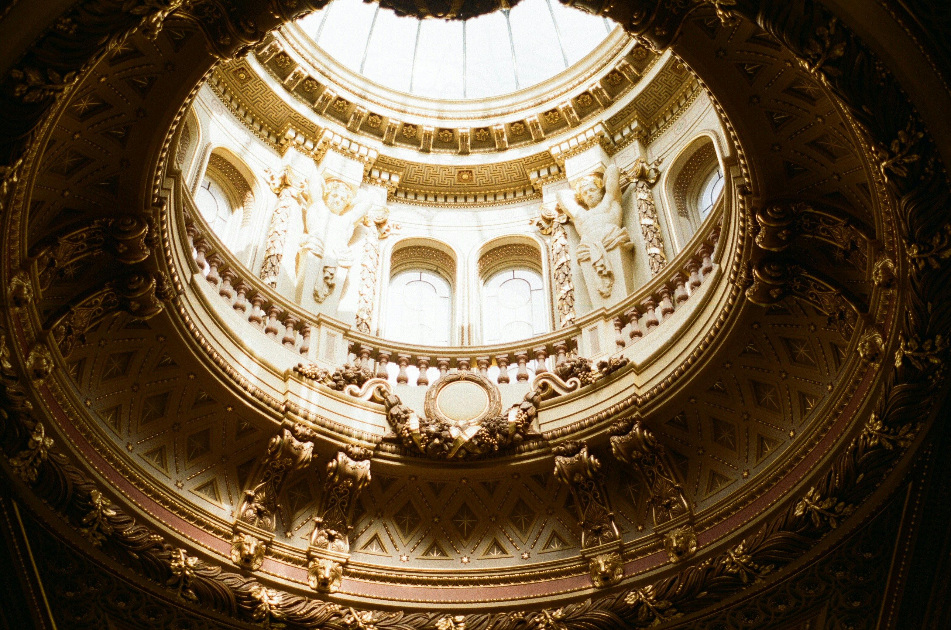 An ornate gilded dome with sculpted figures surrounding a bright circular skylight.