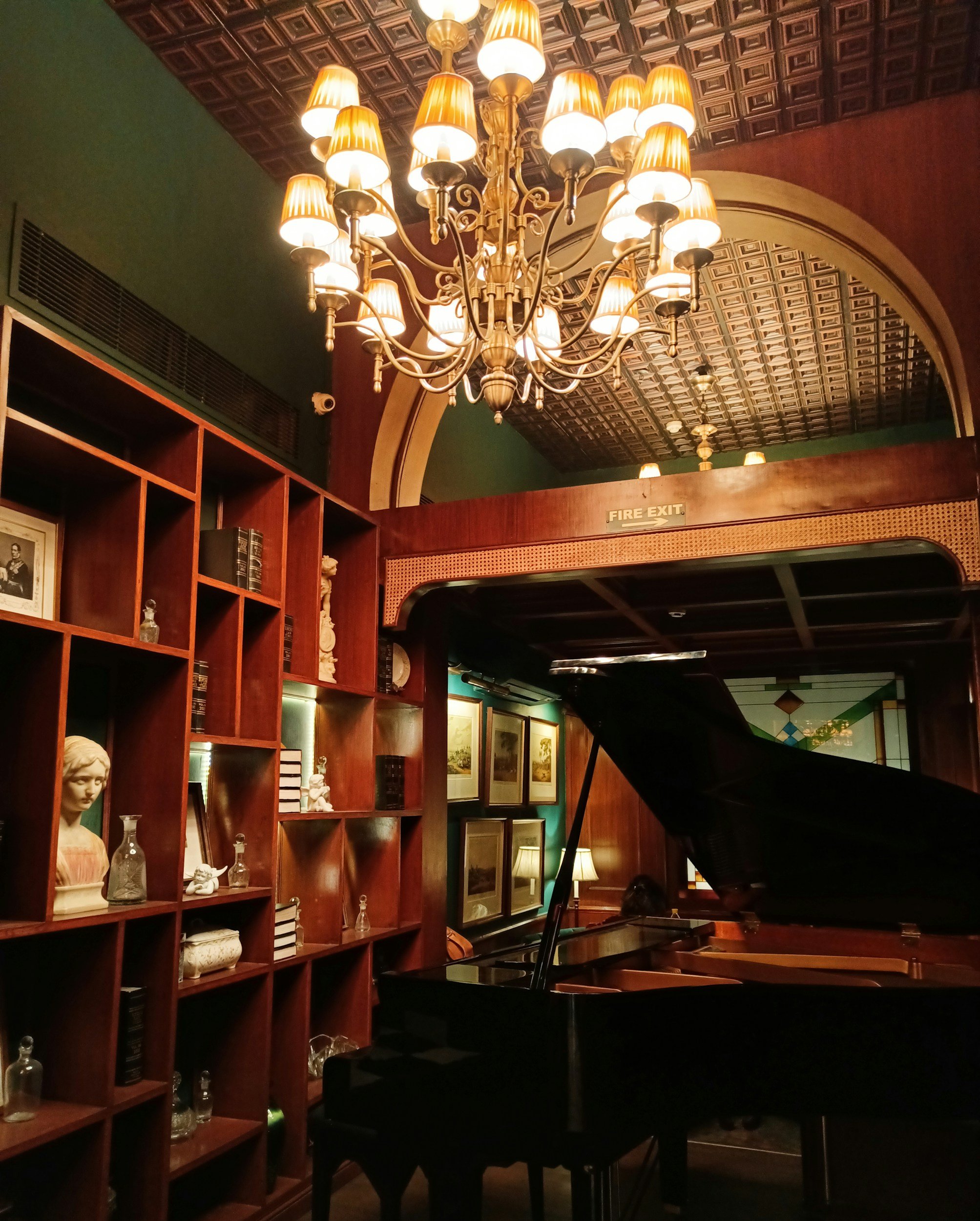 Elegant room with a large chandelier, wooden bookshelves with decorative items, framed artwork, and a grand piano.