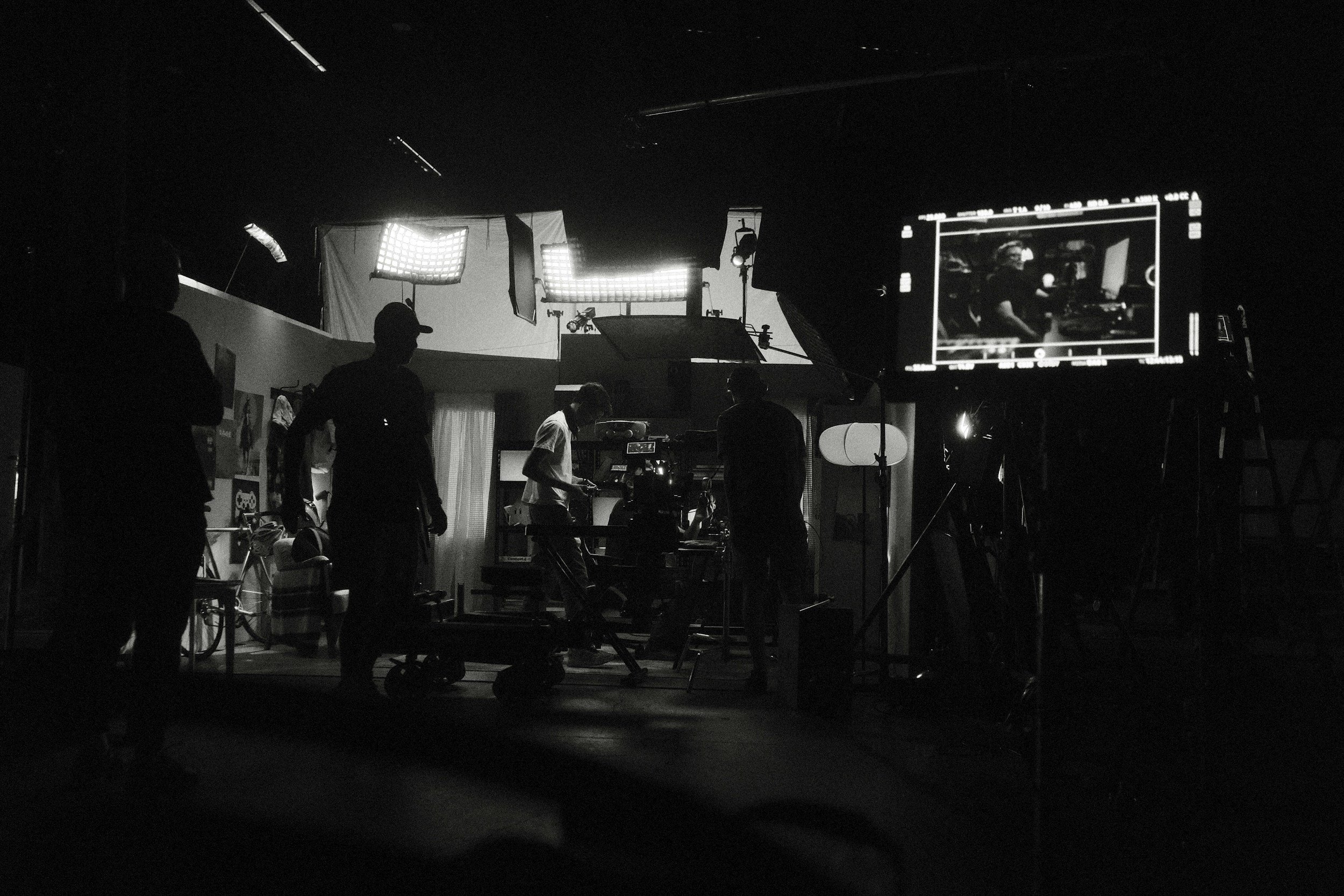 People working on a film set with cameras and lighting equipment, seen in silhouette against a dark background.