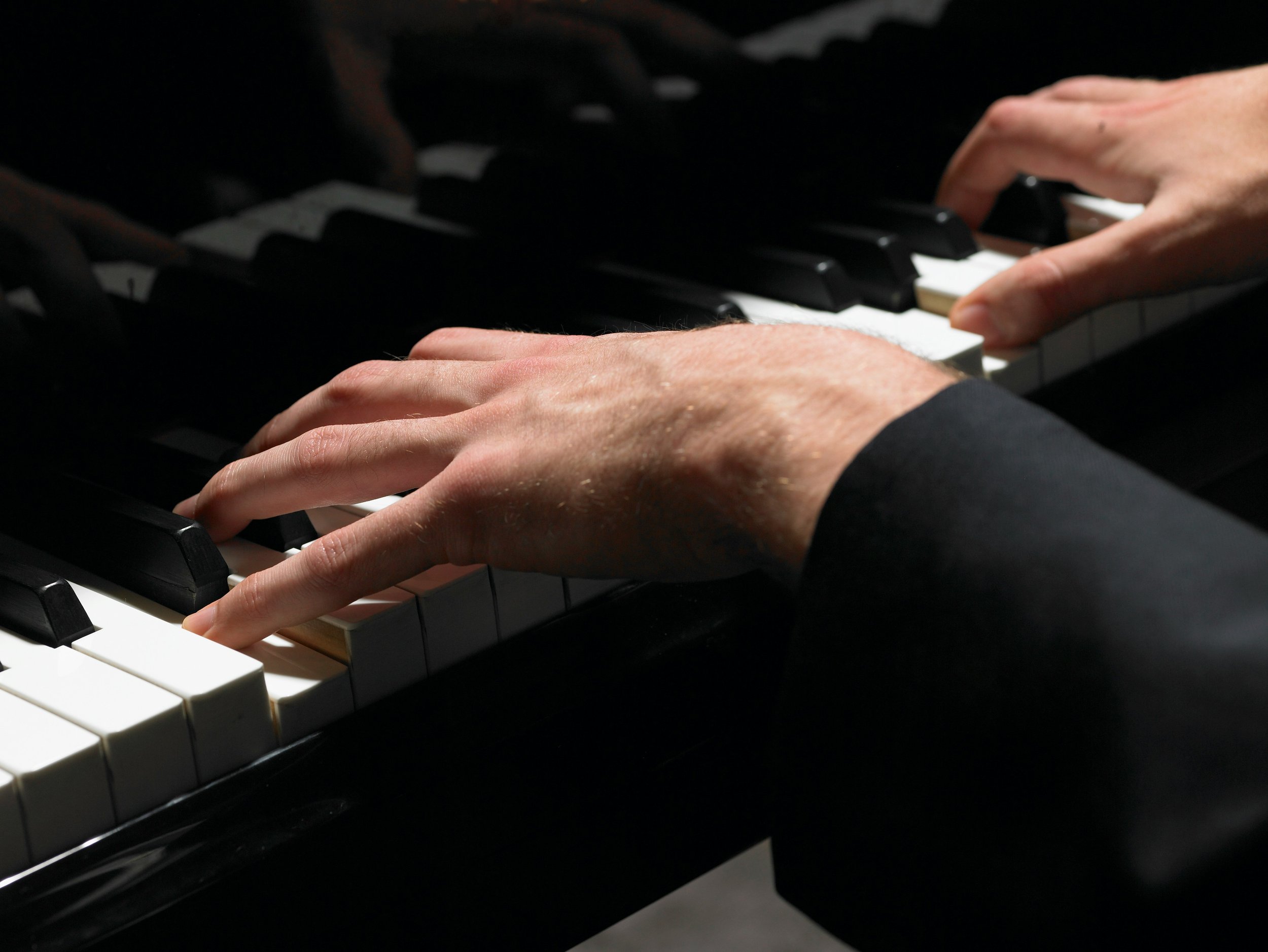 Close-up of hands playing piano, with one hand on black keys and the other on white keys, wearing a black long-sleeve shirt.
